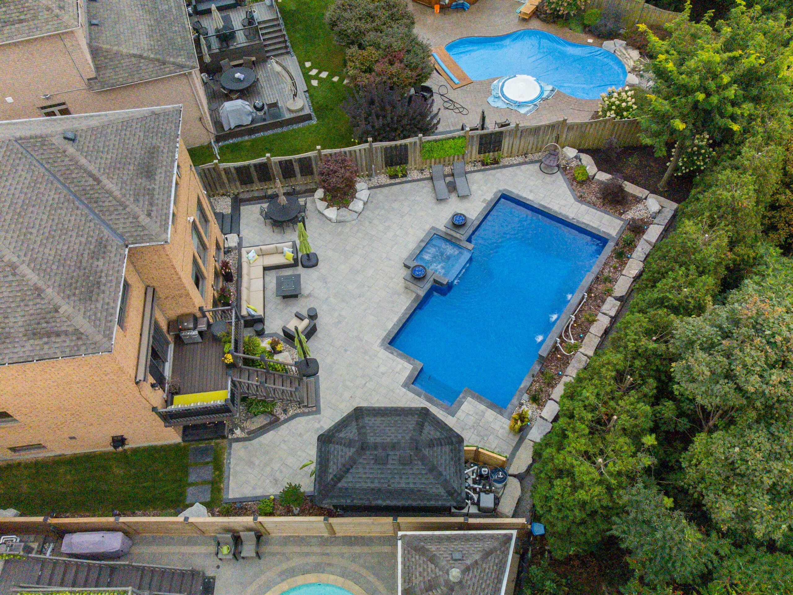 Aerial view of a backyard with a blue swimming pool, patio furniture, and a garden area, surrounded by houses.